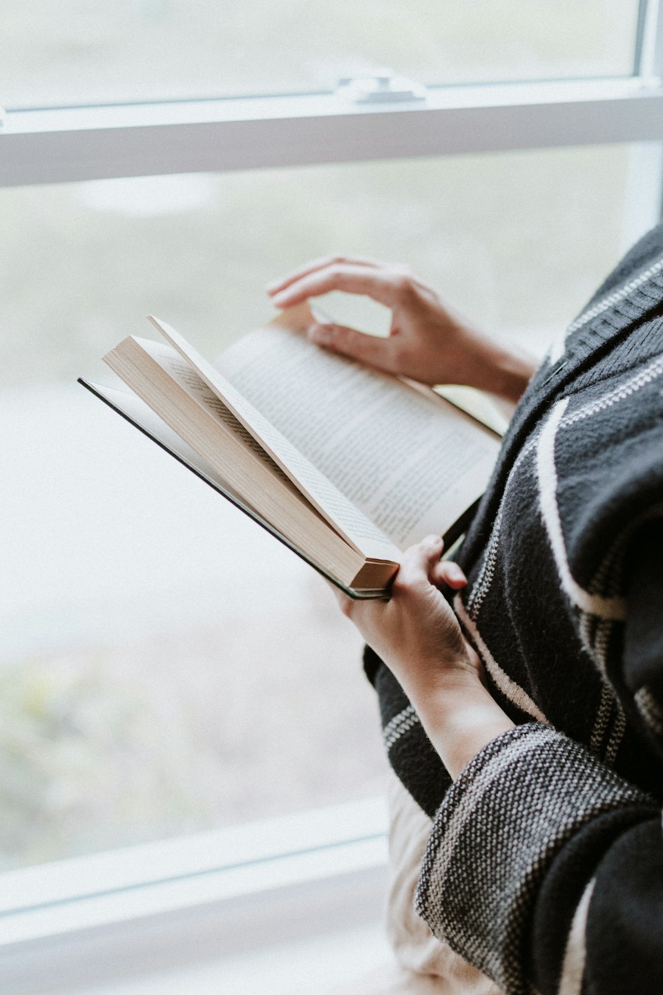 Una persona tiene un libro aperto mentre legge vicino a una finestra.